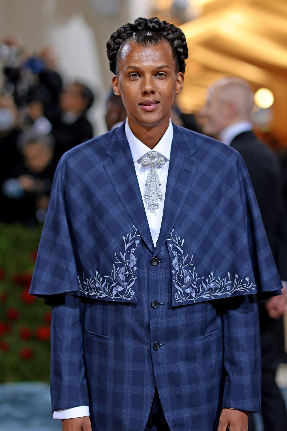 Stromae in a blue checkered plaid suit with an attached cropped cape with additional embroidery. He has a crystal tie on his white button-down. His hair is in a braided updo.