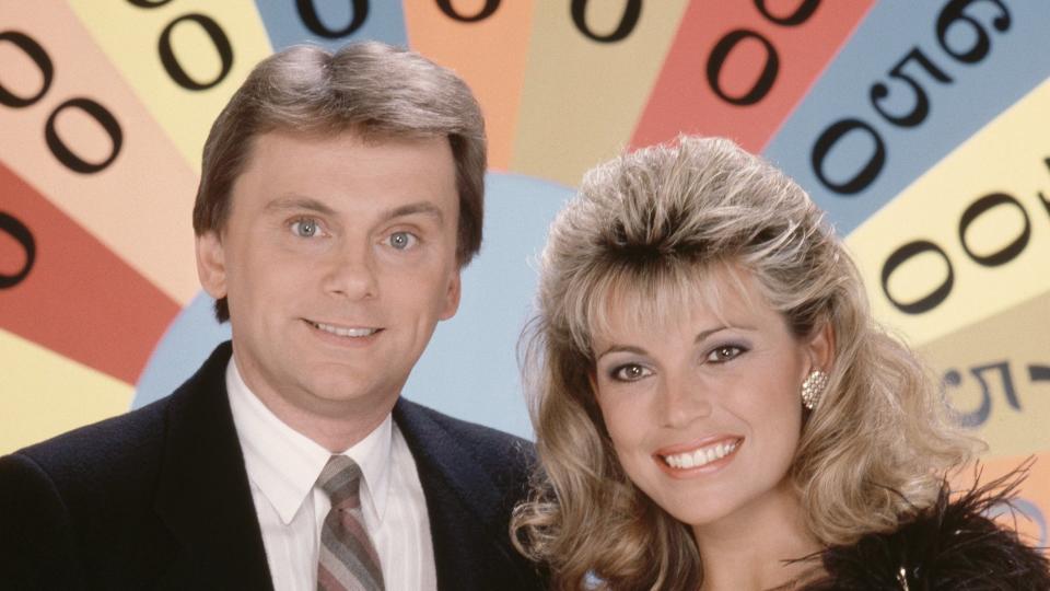 pat sajak and vanna white smile for a posed photo while standing in front of a wheel with money slices, he wears a black suit jacket, white collared shirt, and multicolored tie, she wears a gold glittery one shoulder dress with feathers on the strap