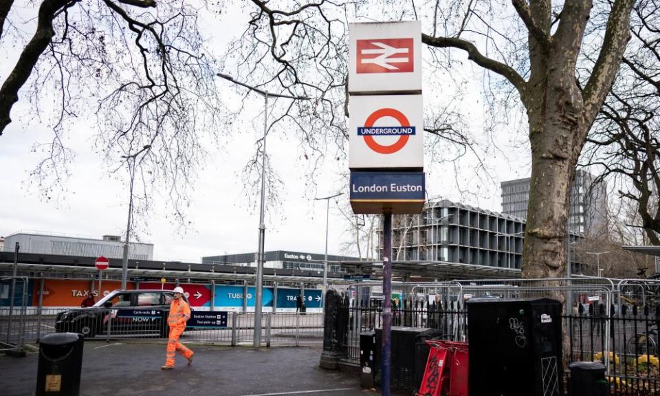 A man in a hi-vis worksuit walking along the pavement of a busy road next to a sign showing the old British Rail symbol and an Underground symbol