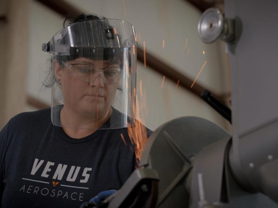 A woman wearing a clear welding mask and perspex goggles while using heavy equipment in a warehouse.