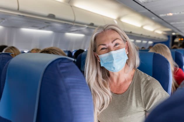 A front-view shot of a mature caucasian woman sitting on an airplane wearing a protective face mask, she is excited to be traveling on holiday. (Photo: SolStock via Getty Images)