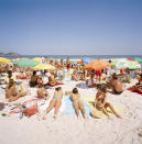 Crowded beach scene with various individuals sunbathing and standing; no recognizable people