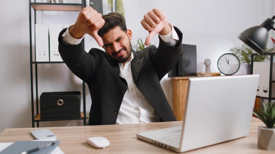 Upset businessman showing thumbs down, sitting at his desk in front of a laptop.