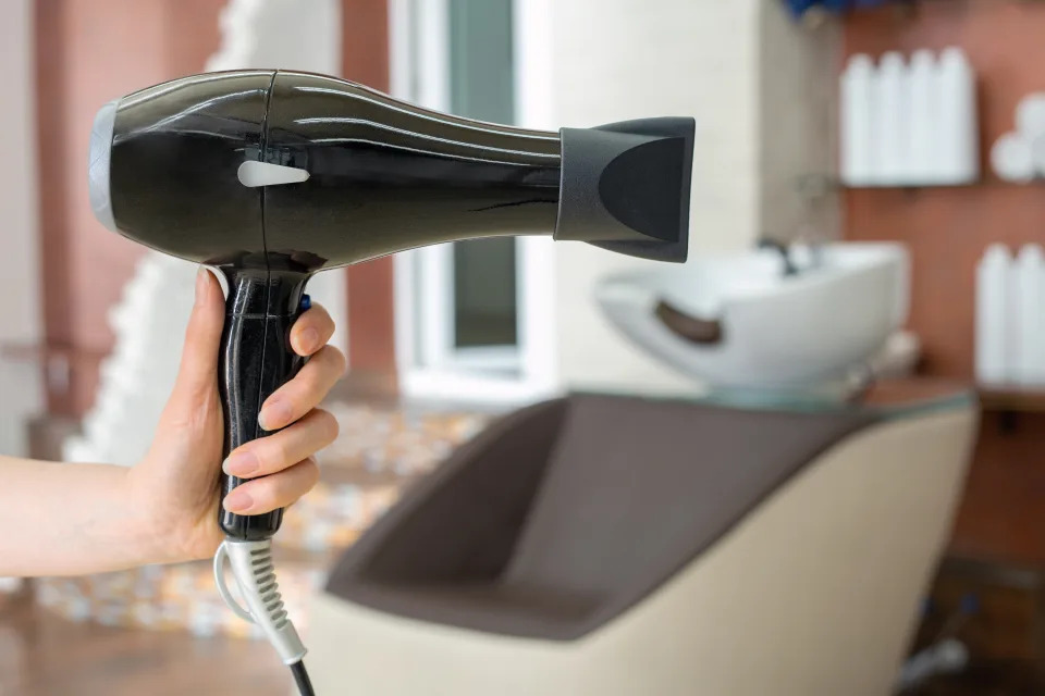 Hair dryer professional hair dresser tool in hairdresser's hand against beauty salon interior background. Hair stylist work space. Beauty hairdressing salon, Barber shop, beauty services for hair.