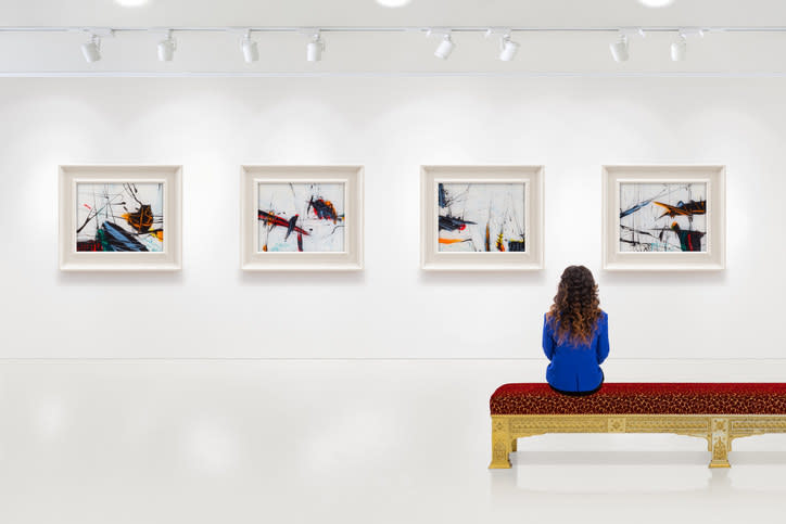 In a exhibition centre, young woman visits an art exhibition and watches artist's collection on the wall. Lightened white wall contains four white frames with artist's painting.
