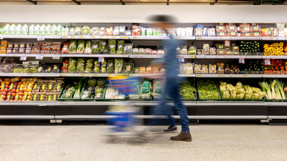 Person pushing a shopping cart by a well-stocked grocery store produce section with various fruits, vegetables, and packaged goods on display