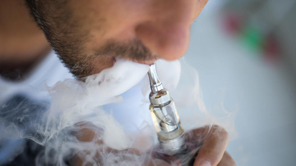 A man smokes an electronic cigarette on a gray background, blowing a stream of smoke.