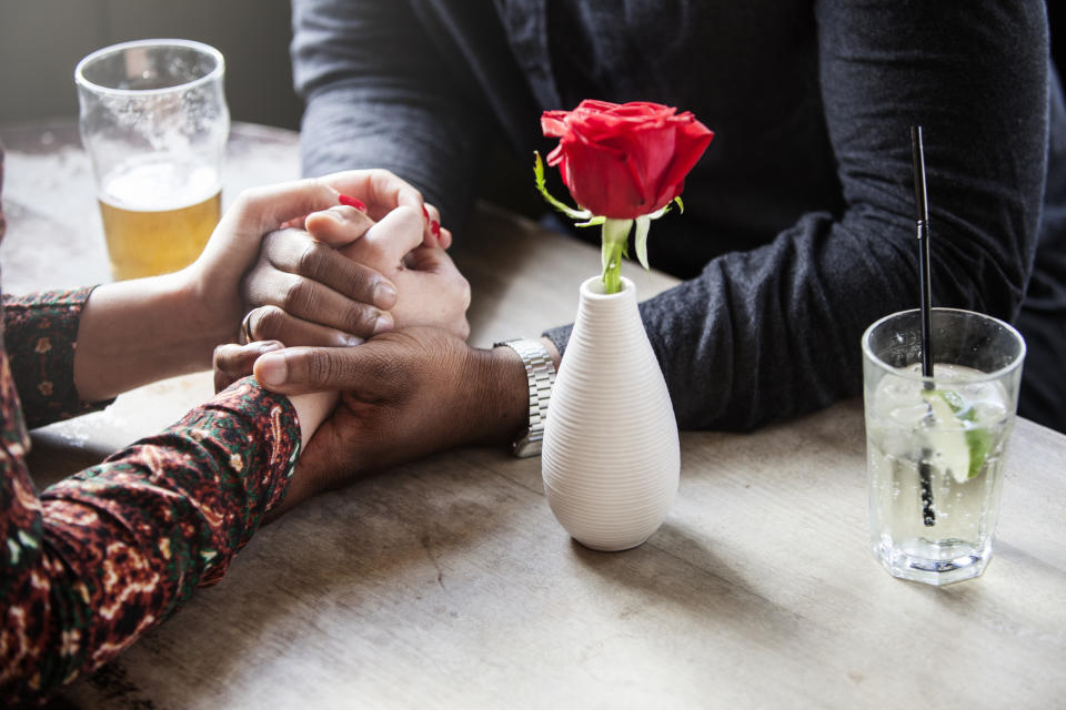 Two people hold hands across a table with drinks and a red rose in a vase between them