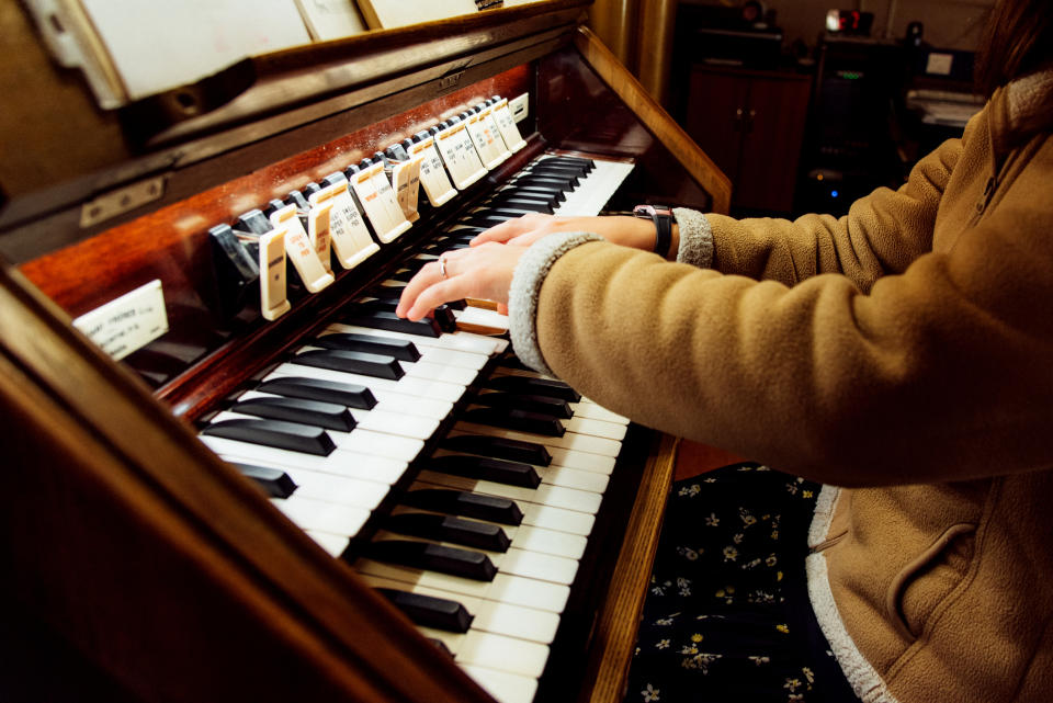 A person in a casual jacket playing an organ. The image focuses on the hands and keyboard