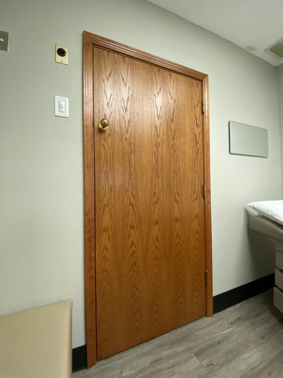 A closed wooden door in a room with a whiteboard and examination bed on the right side, typical of a medical office setting