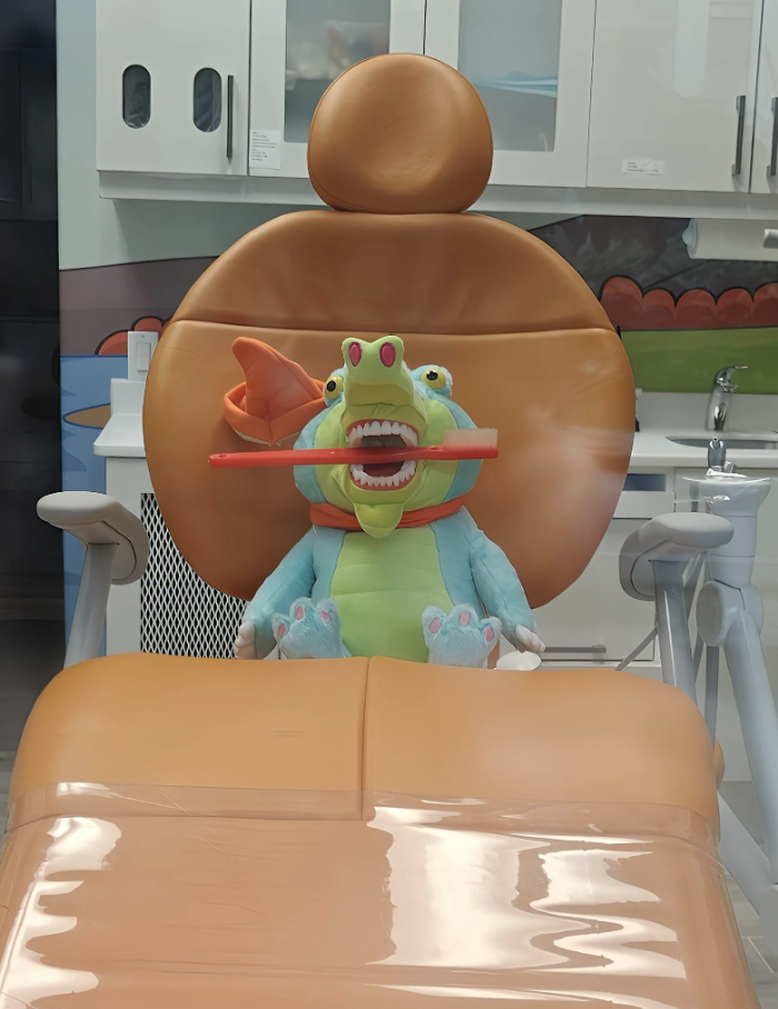 A stuffed toy dragon with a large open mouth sits in a dentist's chair with a toothbrush in its mouth