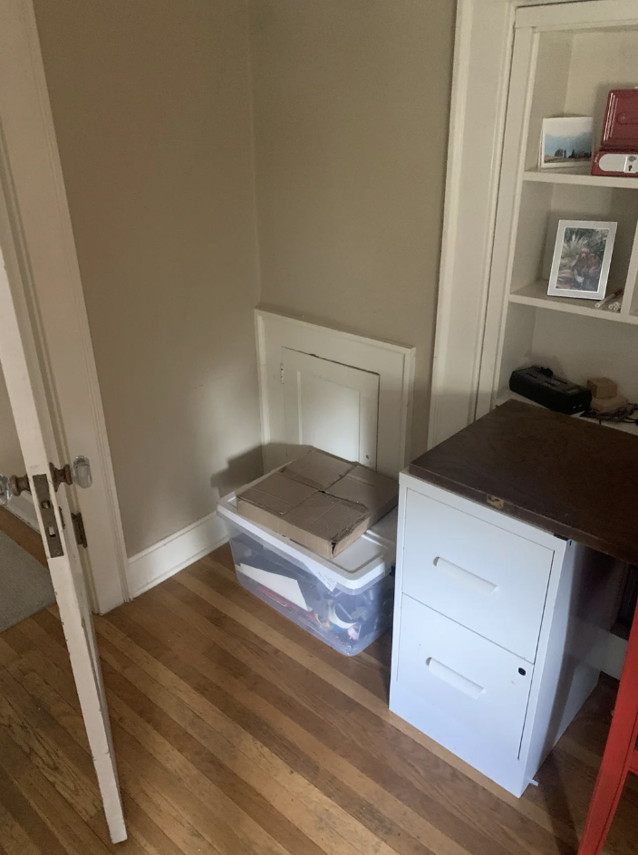 A small, cluttered corner in a home office with a white filing cabinet, a wooden desk, and a plastic storage bin with cardboard on top. Shelves with books and a framed picture are visible