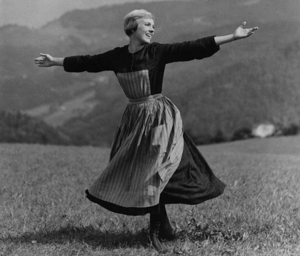 Black and white movie still of a young, short-haired woman frolicking in a field, singing, with her arms spread wide.