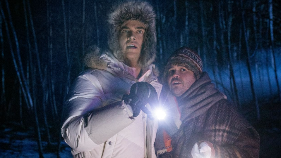 Two men dressed in winter coats look scared in the woods.