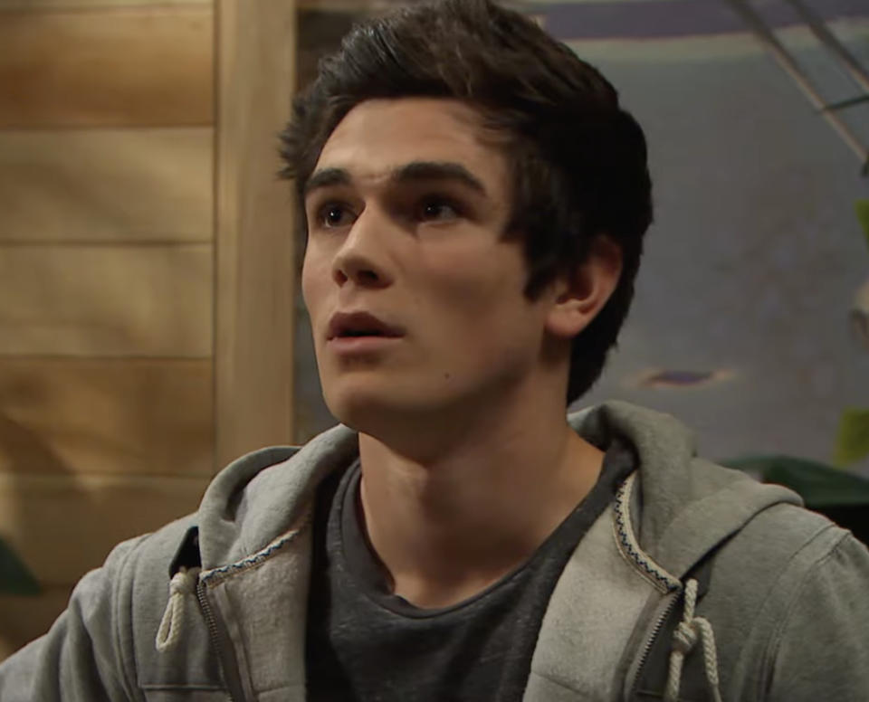 KJ Apa with a surprised expression wearing a gray hoodie in a room with a wood wall background
