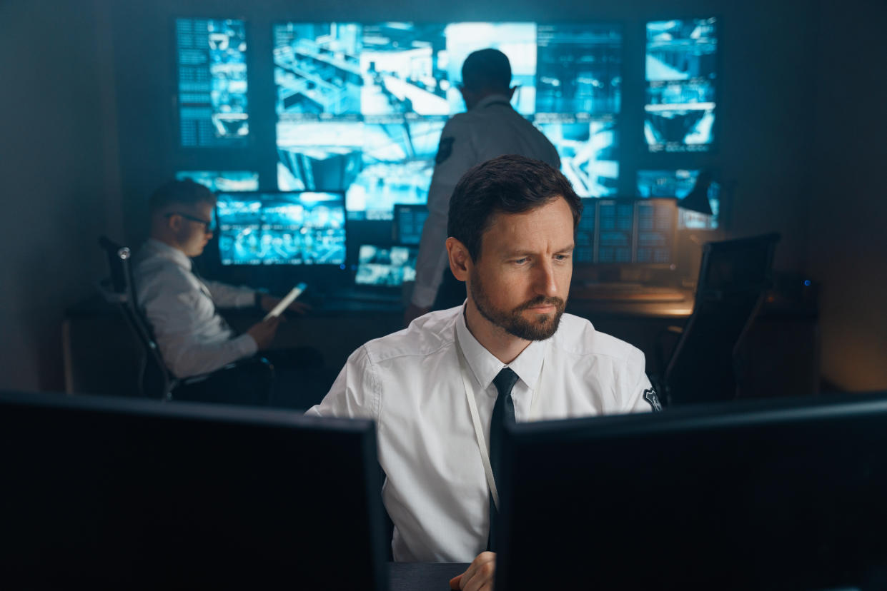 A man sitting at a desk a looking at a monitor with more monitors behind him.