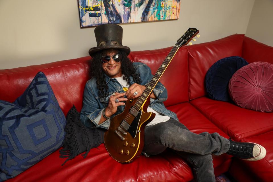 Slash sitting on a red couch with a guitar leaning on his thigh