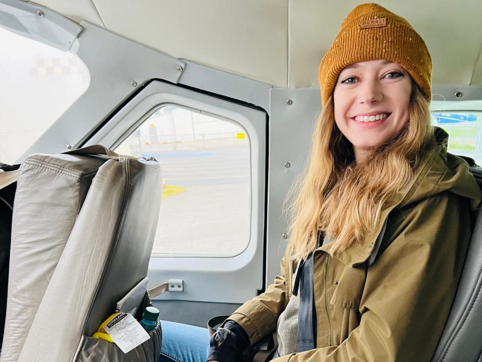 Emily, wearing an orange hat, a beige coat, and blue jeans, smiles while on the plane.