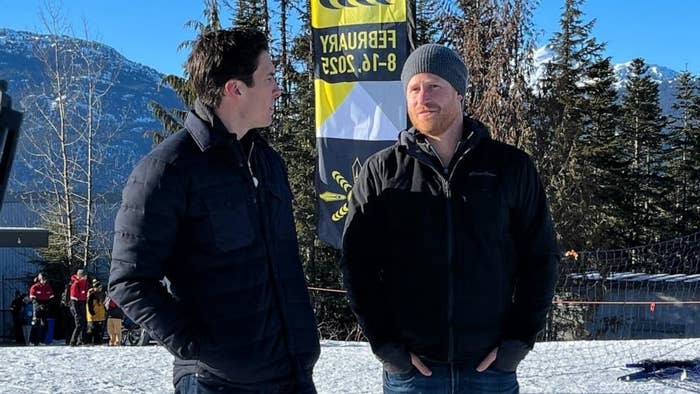 Prince Harry and a GMA anchor in winter jackets stand before a mountain landscape and a "Freewheelers" banner