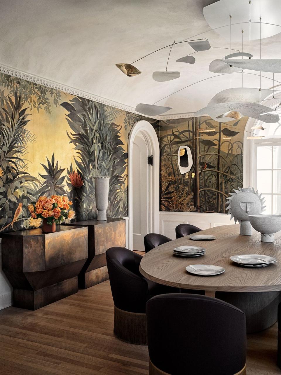 dining room with wooden parquet flooring and bold botanical wallpaper in green and gold tones with an architectural gold wash sideboard and an oval light wash wood table and fringed silk low chairs