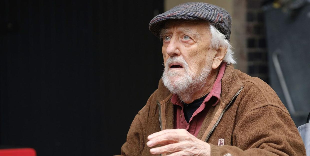 bernard cribbins in doctor who, an older man sits in a wheelchair looking up at someone off camera, he has grey hair and beard and wears a flat cap with brown coat