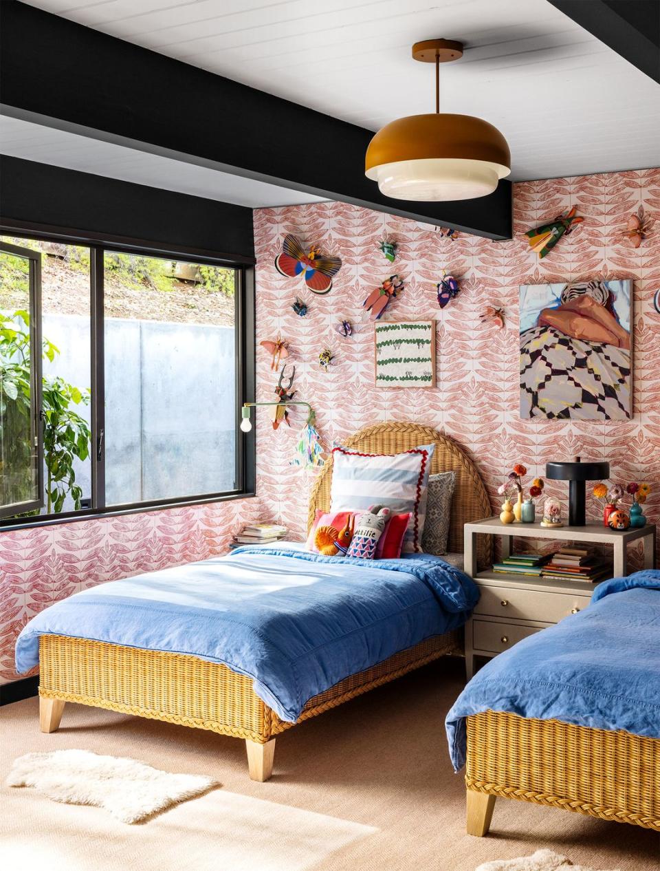 in a kids' room are two twin rattan beds with headboards with blue coverlets and a nightstand between, walls are covered in a leaf patterned wallpaper and decorative butterflies hang on the wall behind the beds