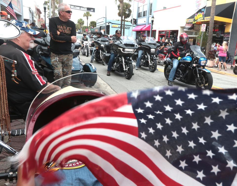 Wind whips an American flag on the back of a parked motorcycle while riders cruise down Main Street on Friday's opening day of Bike Week 2024 in Daytona Beach. "To me, it's like a homecoming," said Johnny Sanchez, owner of John's Rock 'N' Ride, a longtime fixture on Main Street.