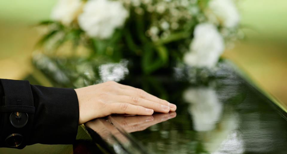 Woman's hand rests of casket with flowers visible int he background.