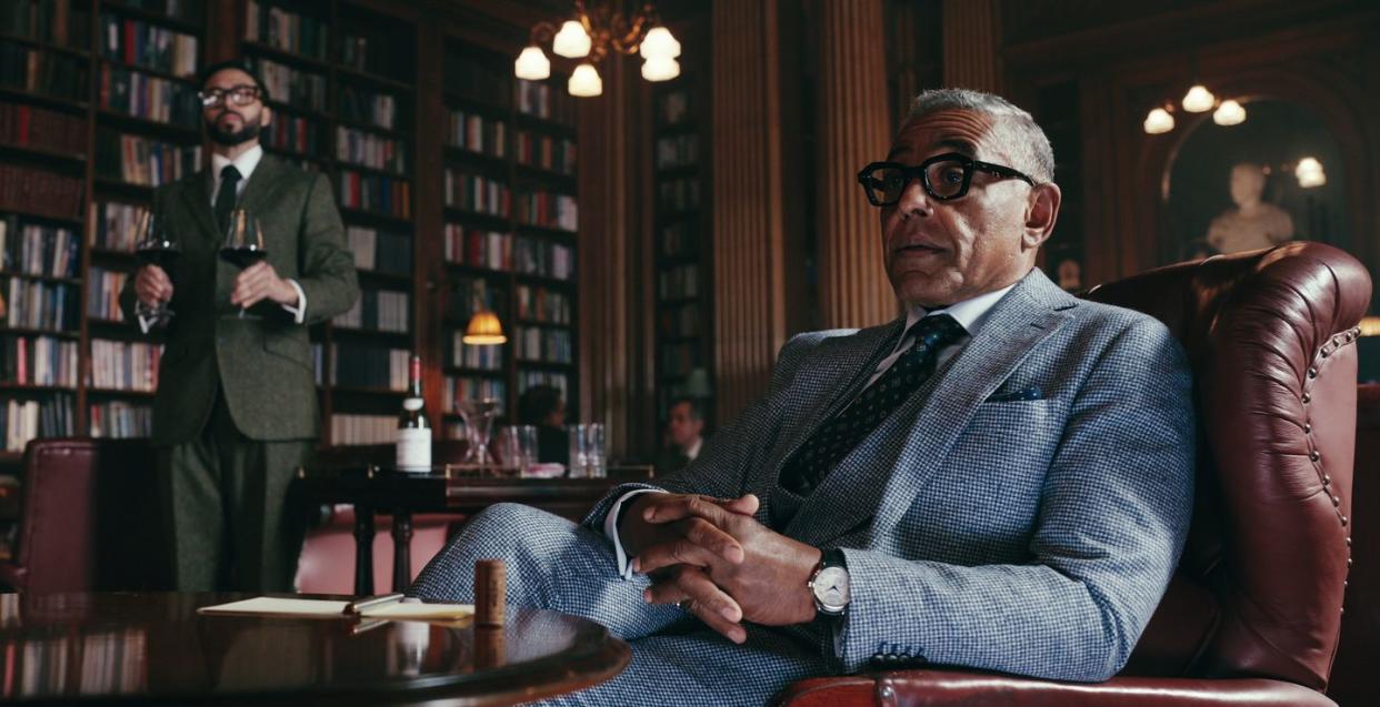 giancarlo esposito in the gentlemen, a man with greying hair and glasses sits in an armchair inside a room with books lining the wall, another man walks up to him holding two glasses of wine
