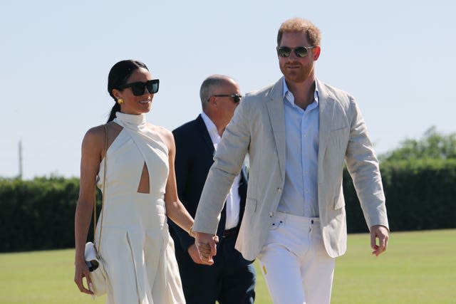 Harry and Meghan, both wearing sunglasses, walking hand in hand