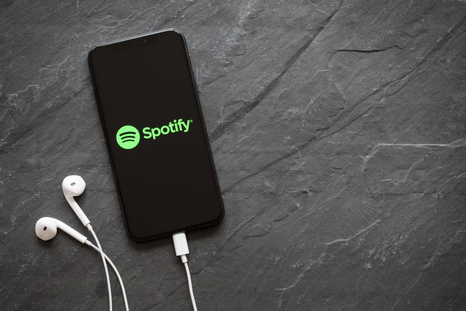 Das Spotify-Logo auf dem Bildschirm eines schwarzen Smartphones, das auf einem graufarbendem Keramikuntergrund liegt. Weiße Kopfhörer sind an das Telefon angeschlossen.
