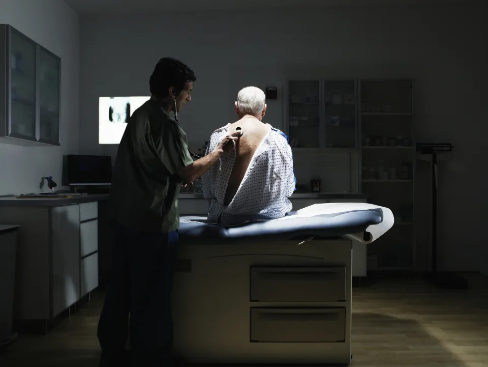 A man in a doctor's appt gown sitting on an exam table while the doctor listens to his heart with a stethoscope