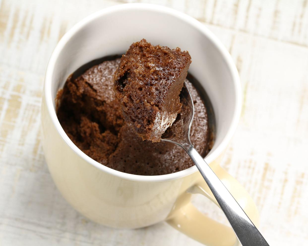 Top-view easy microwave chocolate mug cake in a white porcelain cup, focus on one spoon over the top with a piece of it, on a white painted on beige wooden table