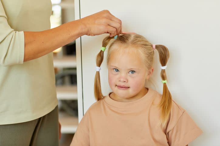 close up of unrecognizable caring mother combing hair of cute girl with down syndrome and tying it in pigtails