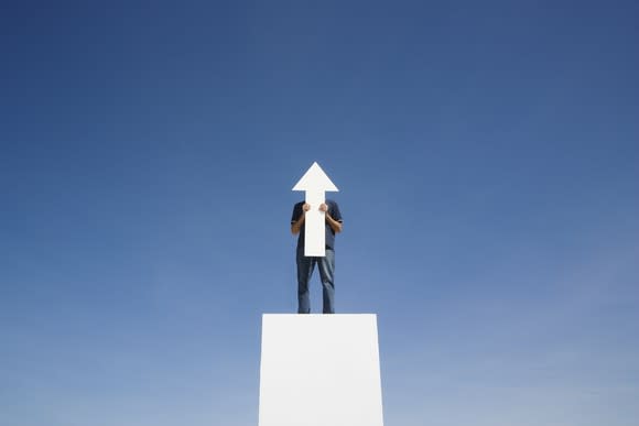 A man standing on a white platform against a blue sky holds an upward-pointing arrow.