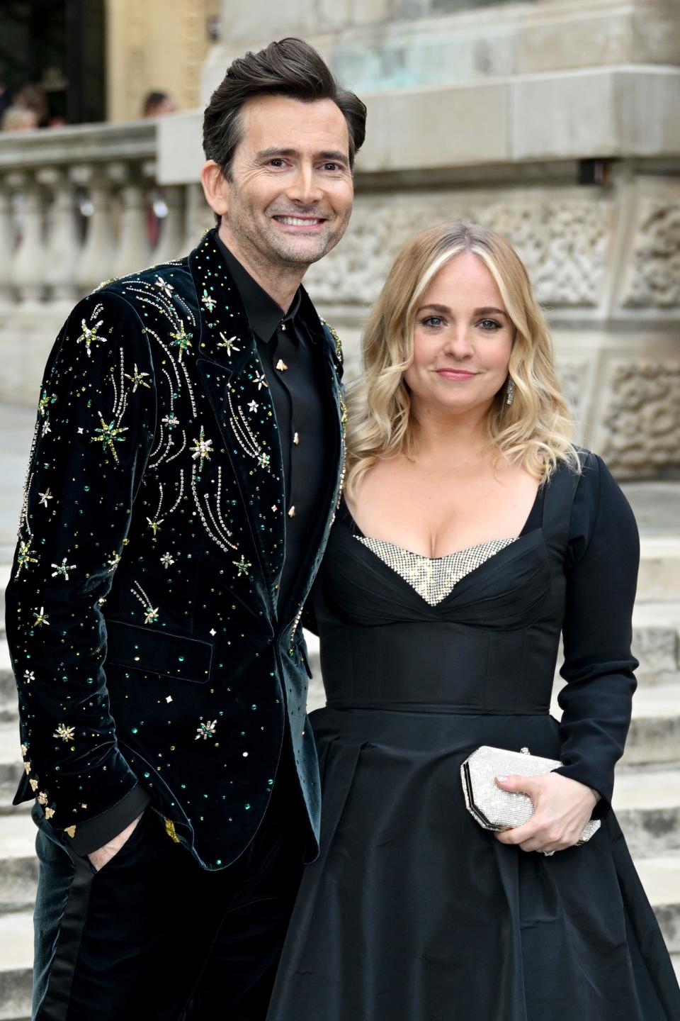 David Tennant and Georgia Tennant pose together on stone steps; David wears a sparkling suit jacket, while Georgia wears a black dress and holds a clutch