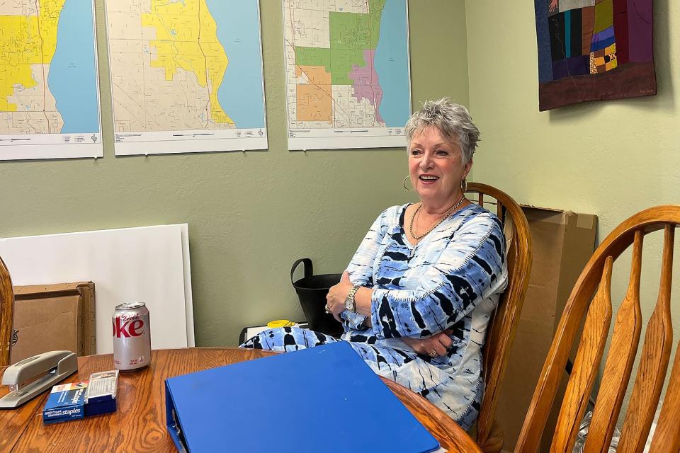 A woman in a blue and white top sitting at a table with her arms folded in a light-green room with maps on the wall.