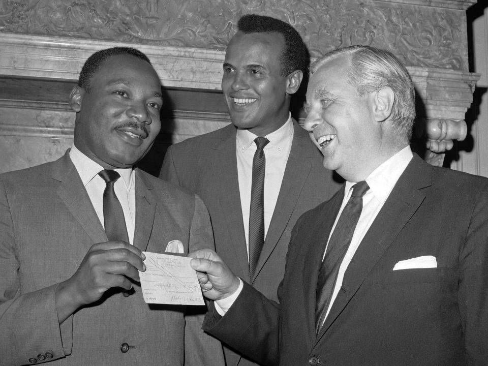 In a black and white photo a man hands another man a check as a man stands behind them with a smile on his face.