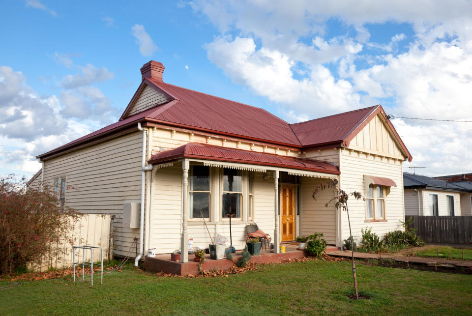 A quaint single-story house with a red roof and pale exterior sits on a grassy lawn under a partly cloudy sky. Gardening tools and plants adorn the front porch