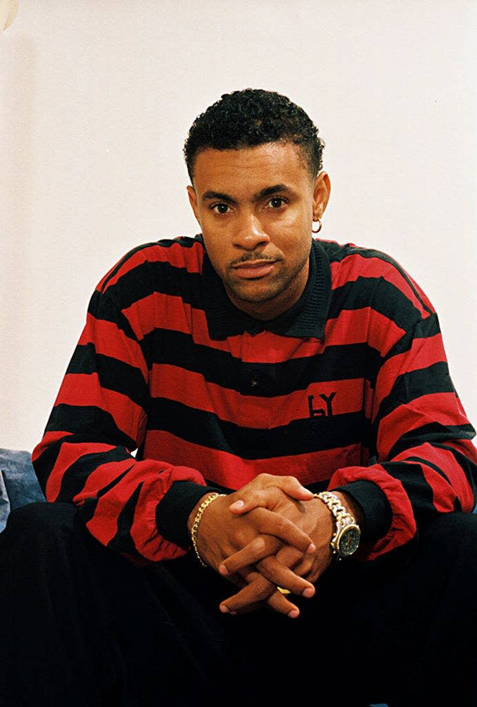 Shaggy looks straight into the camera as he slightly smiles for a photo. He is wearing a striped shirt and a few pieces of jewelry