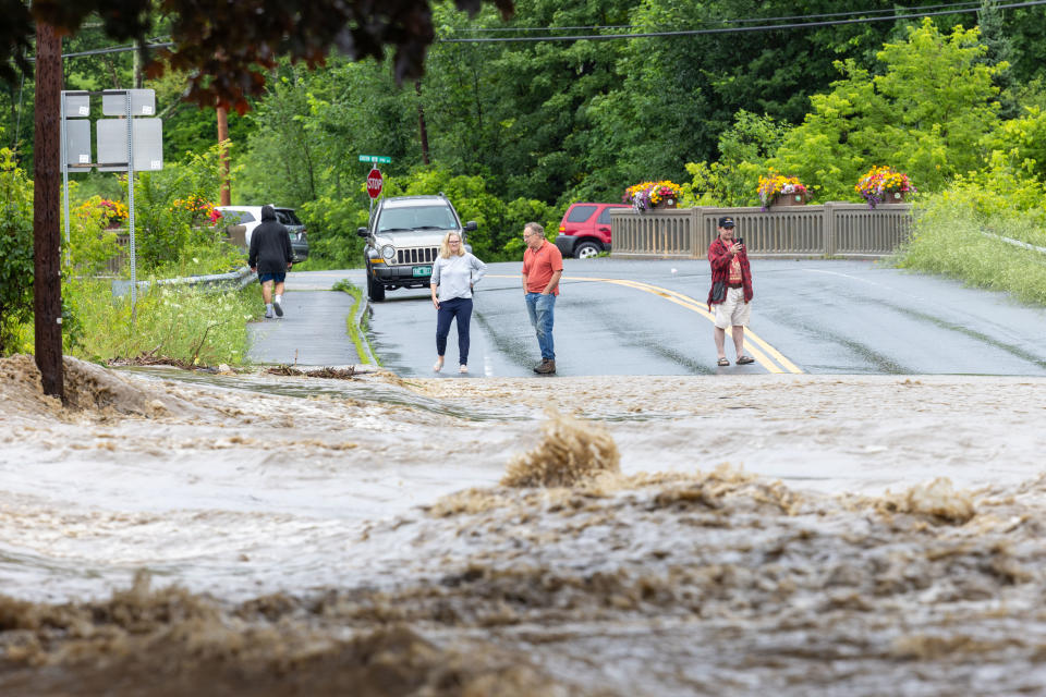 Several people watch as floodwaters triggered by torrential rains flow across a road in front of them.