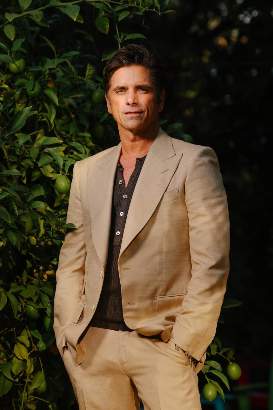 John Stamos in a tan suit and dark shirt stands with his hands in his pockets.