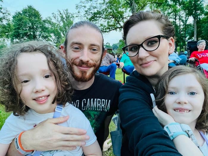 A family portrait of four: two adults, a man and a woman, and two children with curly hair. They are sitting outdoors in a park