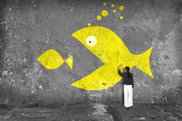 Woman drawing image on a wall of a large yellow fish eating a smaller yellow fish.