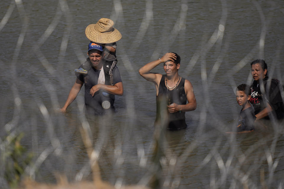 Migrants stand in the Rio Grande behind concertina wire as they try to enter the U.S. from Mexico near the site where workers are assembling large buoys to be used as a border barrier in Eagle Pass, Texas, Tuesday, July 11, 2023. The floating barrier is being deployed in an effort to block migrants from entering Texas from Mexico. (AP Photo/Eric Gay)