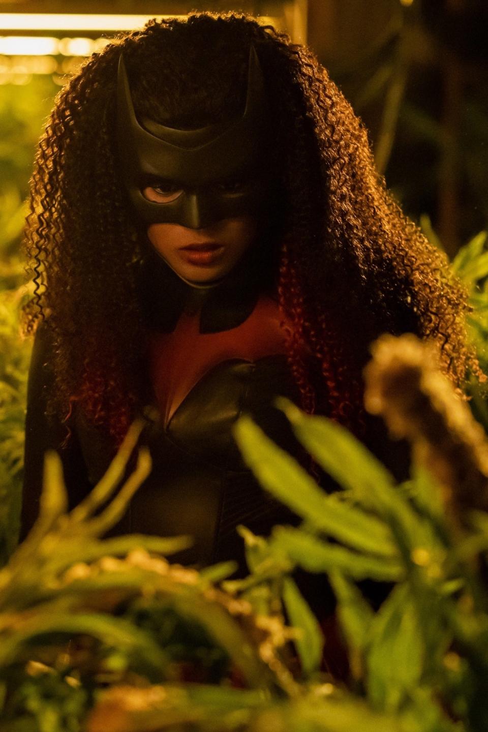 Javicia Leslie dressed as Batwoman stands surrounded by lush foliage. She is wearing a superhero costume with a mask, peering intensely through the plants