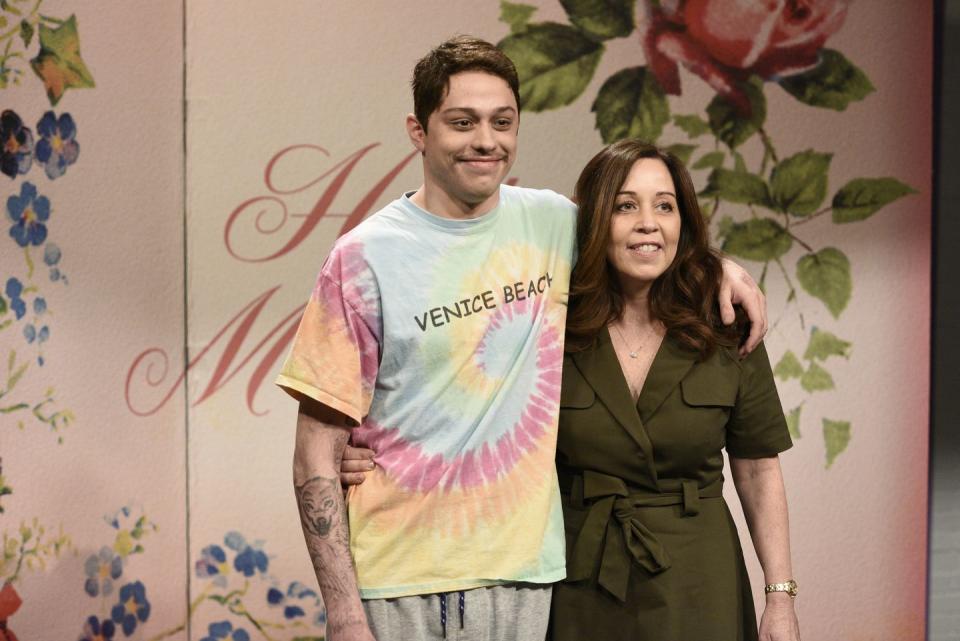 pete davidson and amy waters davidson stand next to each other and smile past the camera, pete has his left arm around his mothers shoulders, amy has her right arm around petes waist, he wears a tide tshirt with the words venice beach across the chest, she wears an olive dress