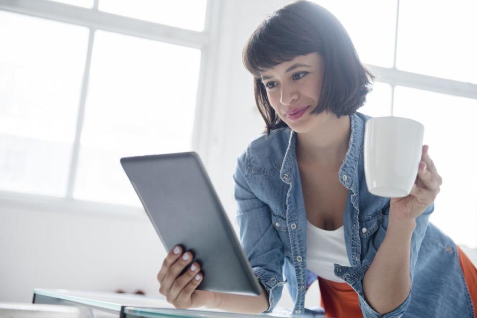 A woman drinks out of a mug while using a tablet