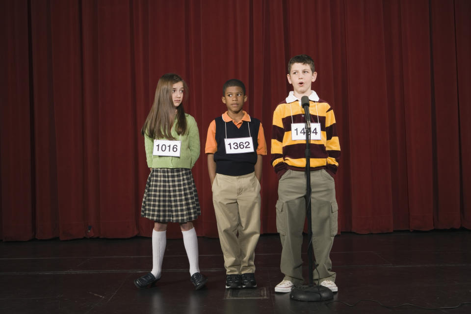 Three children, identified by numbers 1016, 1362, and 1444, stand in a line on stage for a spelling bee competition, with one child speaking into a microphone