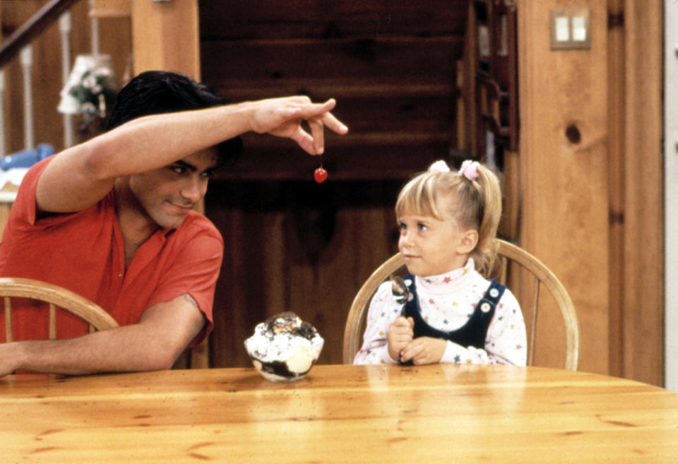 Uncle Jesse and Michelle from Full House sit at a table, with Jesse playfully dangling a cherry over Michelle's head